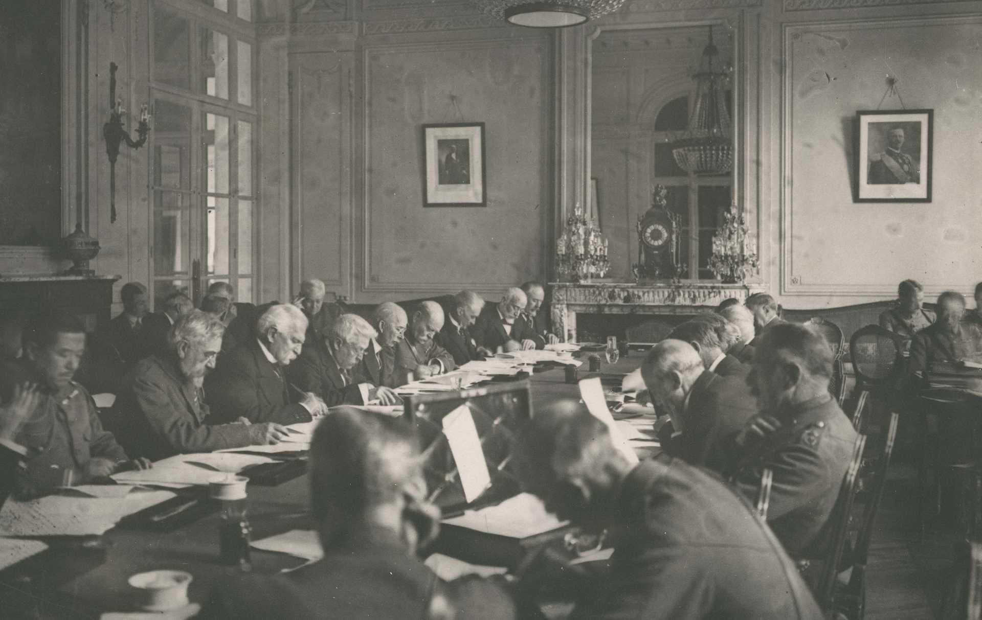 Black and white photograph shows the interior of a large room with members of a committee reading documents at a large table.