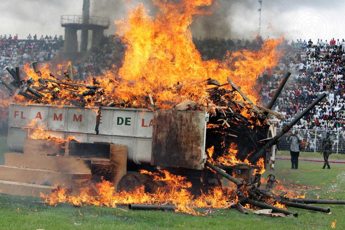 Large crowd in a sports stadium watches a burning container filled with light weapons.