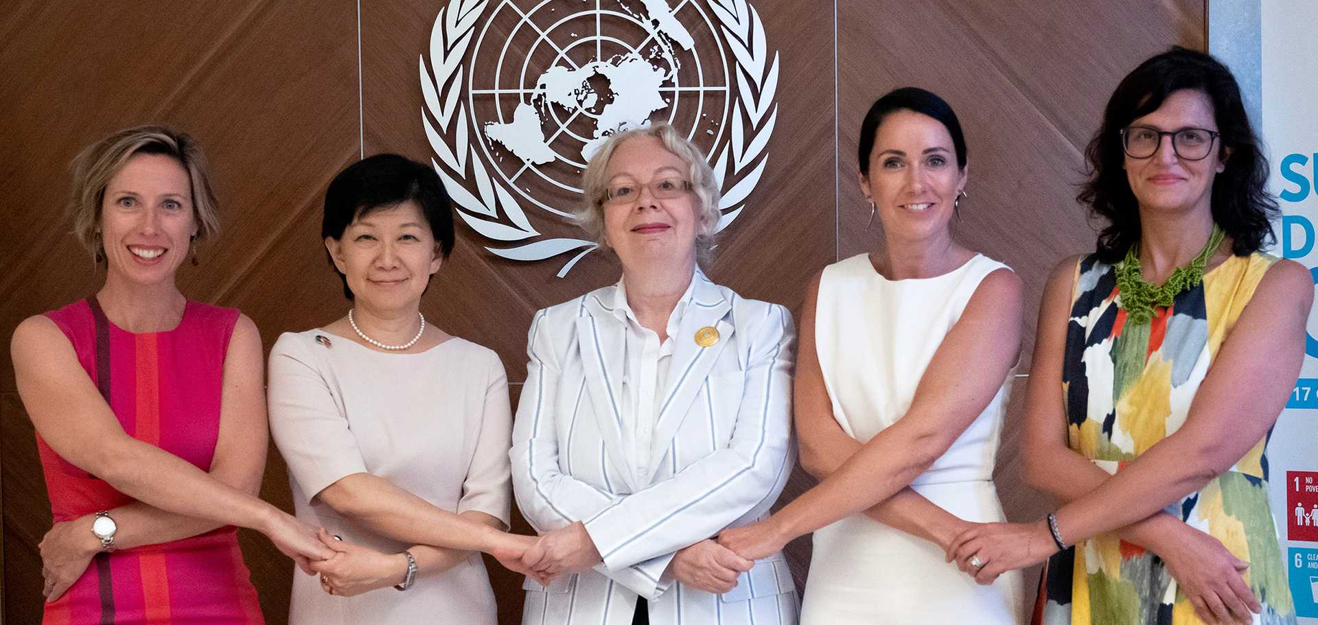 Photo of five women holding her arms crossed and each others hands. On the wall behind them the logo of the UN.